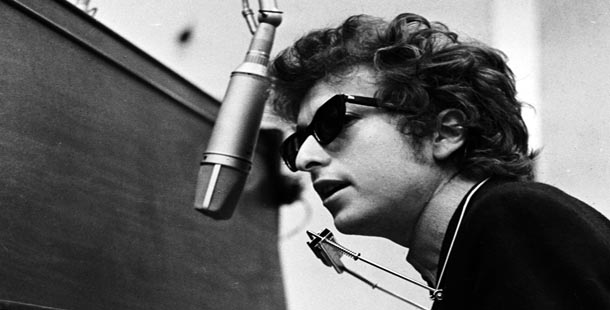 Bob Dylan singing at a mic, neck forward with harmonica attached to his neck